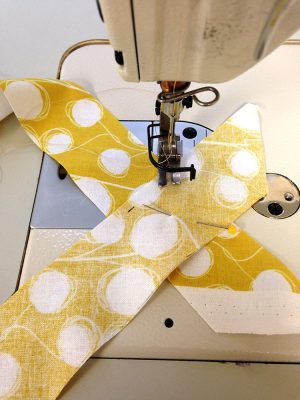A close-up of the needle of a sewing machine, where two strips of yellow fabric patterned with white dots are ready to sew