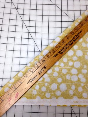 Yellow fabric with white dots, folded diagonally on a cutting board, with a ruler placed on top